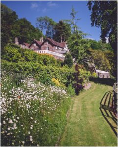 Historical house with red roof in greenery, garden path with flowers, sunny blue sky background.