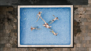 Top-down view of five people in swimwear relaxing in a rectangular pool on a tiled patio.