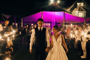 Couple walks hand in hand through sparkler-holding crowd under purple-lit evening tent.