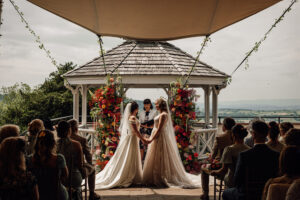 couple weds under floral gazebo, officiant and guests attending, scenic backdrop.