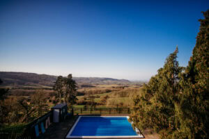 Swimming pool surrounded by greenery with rolling hills and clear blue sky in the background.