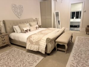 Well-appointed beige bedroom, upholstered bed, decorative heart wall piece, open door to bathroom.