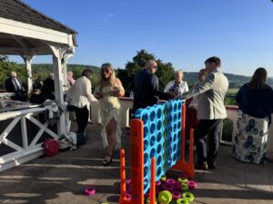 People outdoors playing Connect Four on a patio with green hills in the background.