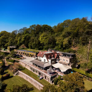 Large hillside building with courtyard, canopy, lush greenery, and forested background.
