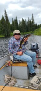 Person in plaid shirt on boat holding fish, forest and cloudy sky in background.