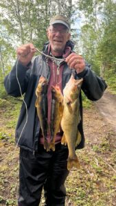Adult in forest holding three fish on a stringer, wearing cap, glasses, and outdoor clothes.
