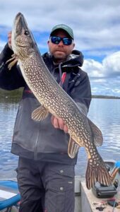 Person on a boat holds large fish under cloudy sky, wearing sunglasses and cap near water.
