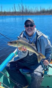 Person fishing on a lake, holding a fish, wearing sunglasses and a cap with clear sky.