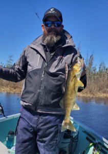 Person on boat with jacket and sunglasses holding fish, trees and blue sky in background.
