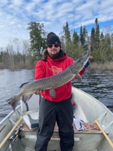 Person in red sweatshirt holding large fish on boat in lake surrounded by trees, cloudy sky above.