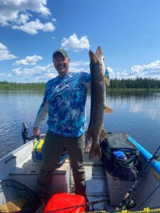 Person on a boat holds large fish, wearing fishing shirt and cap, lake and forest in background.