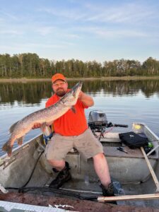 Person in orange shirt holding large fish in boat on calm lake, tree-lined shore, blue sky.