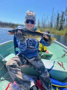 Person in a boat wearing blue hoodie, camouflage pants, sunglasses, holding a fish, lake view.