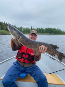 Person in boat holding large fish, wearing red life jacket and camo hat, lake and forest behind.