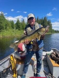 Person on boat holding large fish in forested water setting under clear sky.