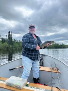 Person on a boat in a forested lake holding a fish; overcast sky with fishing gear on board.