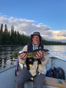 Person in a boat on a lake, holding a fish, with forest and fishing gear visible.