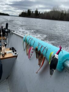 Colorful fishing lures on blue holder in boat, moving on water with rods and trees visible.