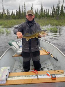 Person in boat fishing, holds large fish, wearing waterproof gear; forest visible in background.