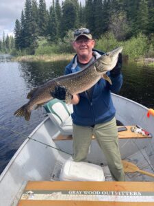 Man in boat on lake holding large fish, evergreen trees in background, fishing gear visible.