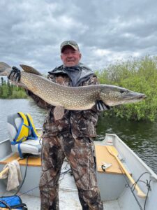 Person in camouflage on boat holding a northern pike with cloudy sky and wooded background.