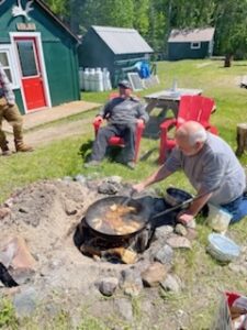 Two people outside small cabins; one cooking on open fire, one seated in a red chair.