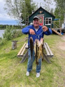Person holding fish by a cabin on grassy lakeside area with trees and partly cloudy sky.