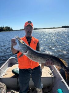 Man in orange vest and hat holds large fish in boat on a calm lake with trees and blue sky.