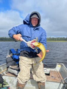 Person in blue jacket holds a fish on a boat; cloudy sky and forested shoreline in background.