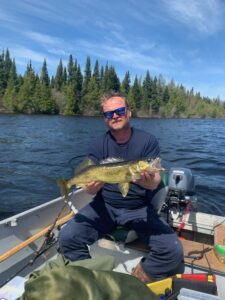 Man in boat on lake holds large fish, with forest and blue sky. Fishing gear visible.