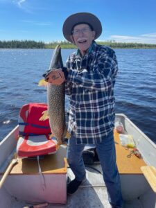 Elderly person in a boat holds a large fish on a sunny lake, wearing plaid, jeans, hat, gloves.
