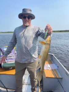 Person on a boat holding a caught fish with fishing gear and treeline in the background.