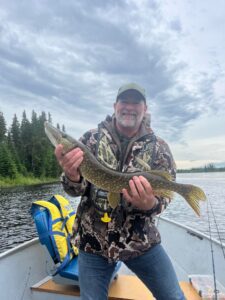 Person in camouflage with fish on boat, lake and forest in background, overcast sky.