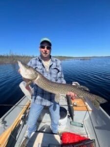Person on boat holding large fish on lake with blue sky and trees in background.