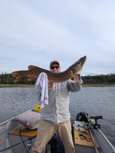 Person on boat holding large fish with cloth, water and wooded shore in background, fishing gear visible.