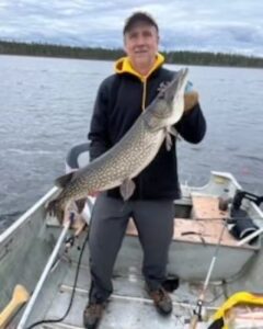 Person standing in boat on lake holding large fish, fishing gear visible, cloudy sky, tree-lined shore.