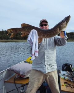Person on boat holds large fish with towel, fishing gear and trees in background.