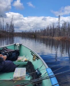 Person sitting in a fishing boat on a calm lake, surrounded by bare trees and cloudy sky.