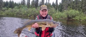 Person holding large fish in a boat on a river, wearing red life jacket and hat, trees behind.