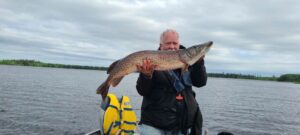 Person on boat holding large fish, life jacket in foreground, cloudy sky, distant trees.