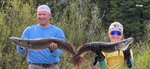 Man and child outdoors holding large fish, surrounded by greenery.