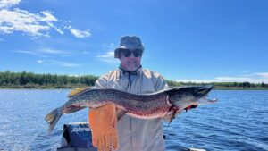 Person in sunglasses holds large fish on a boat, sunny lake, trees, clear sky.