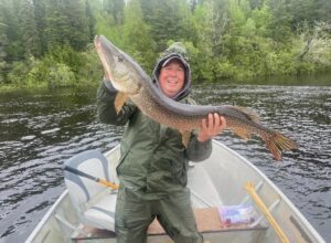 Person in rain gear on a boat holding a large fish over forested water.