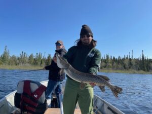 Two people on a boat; one holds a large fish. Forested background under a blue sky.