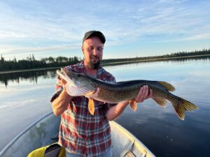 Man in plaid shirt holds large fish on boat with calm lake and tree-lined shore in background.