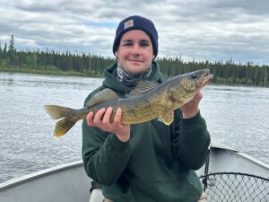 Person in green hoodie and beanie holds a fish on a boat with forest and cloudy sky backdrop.