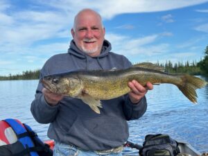 Man on boat holding a large fish with lake, trees, and clear blue sky in the background.