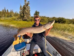 Person in sunglasses holding large fish on boat in lake, trees in background, clear sky above.