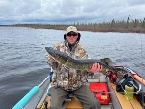 Person in camo with sunglasses holding a large fish on a lake boat, cloudy sky, treeline behind.