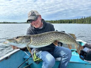 Person on boat holding large northern pike; forested shoreline in background.